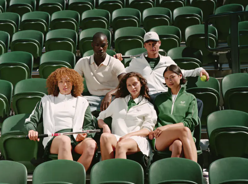 Five diverse young athletes in green and white tennis outfits pose with rackets and a ball in empty green stadium seats