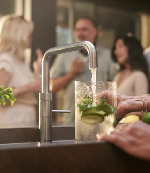 Hands holding a glass of infused water with cucumber and mint under a modern kitchen faucet, people socializing in background