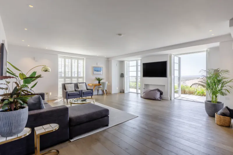 Modern open-plan living room with grey sofas, wall-mounted TV, fireplace, and large glass doors opening to a scenic coastal view