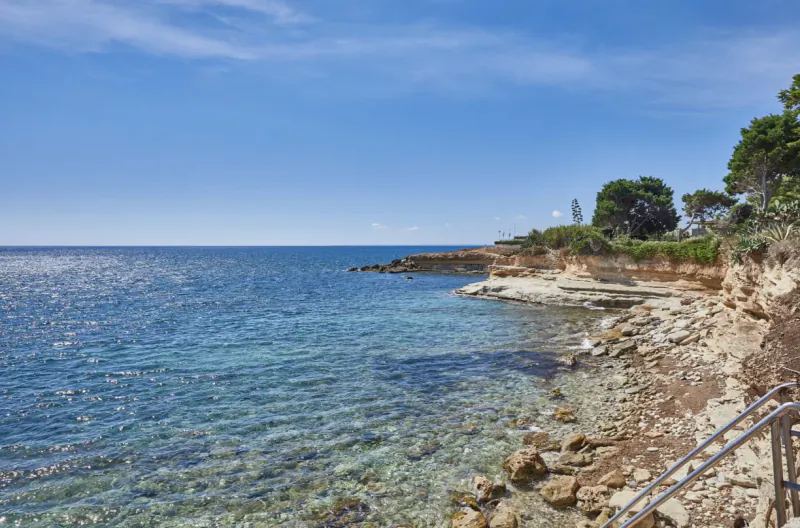 Clear blue sea with sparkling sunlight reflecting on the water, rocky coastline and cliffs with green trees under a bright sky