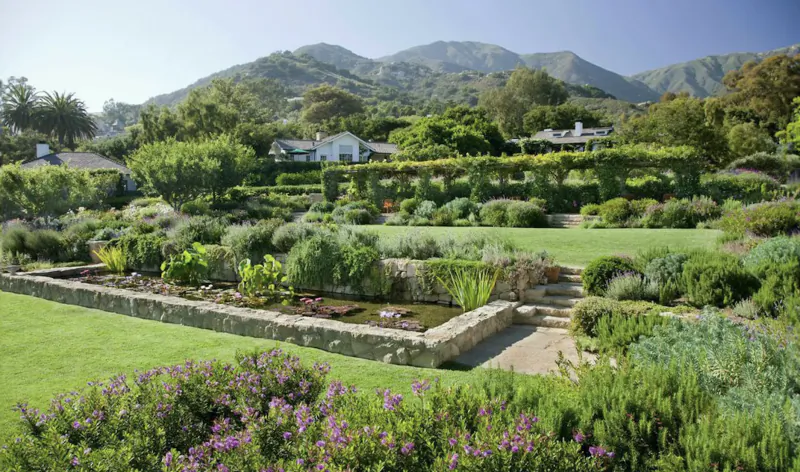 Lush hillside garden with stone pond, terraced greenery, and mountain backdrop