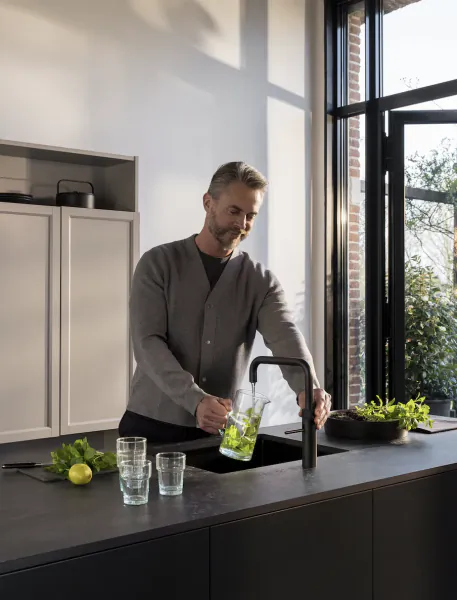 Man filling a glass pitcher with water from a modern black kitchen faucet