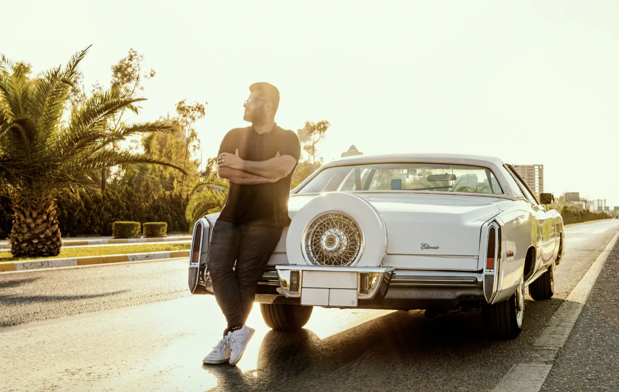 Man in black t-shirt leaning against the rear of a white classic car on a sunny road with palm trees