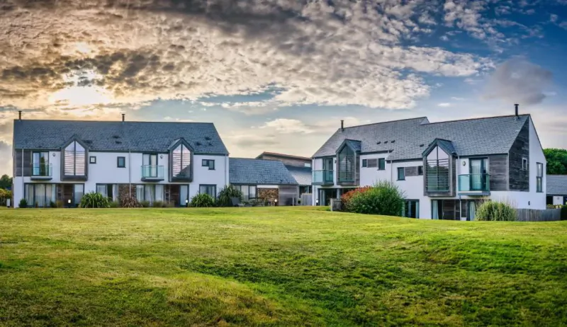 Modern white and grey holiday homes with large glass windows overlooking a green lawn at sunset