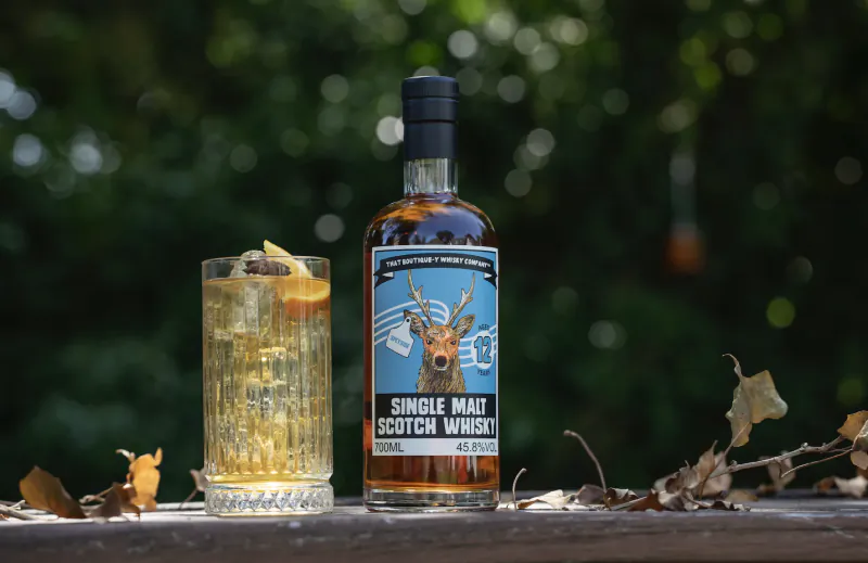 Bottle of single malt Scotch whisky and a glass with ice and orange peel on a wooden surface outdoors