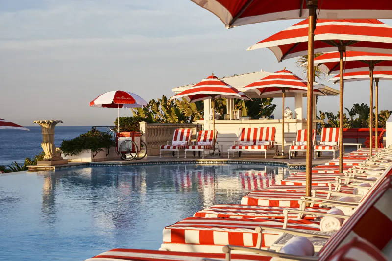 Luxury poolside with red and white striped lounge chairs and umbrellas overlooking the ocean at sunset