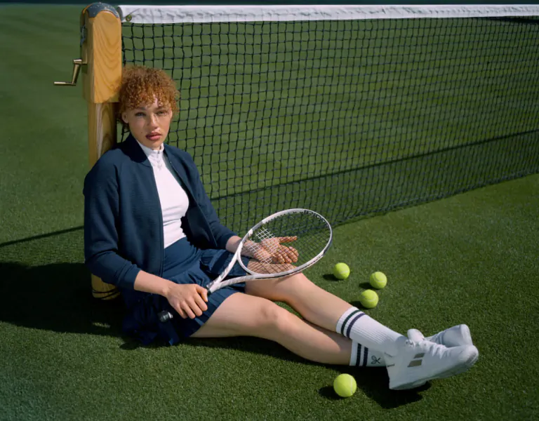Woman in navy tennis outfit sitting on grass by net, holding racket with tennis balls nearby