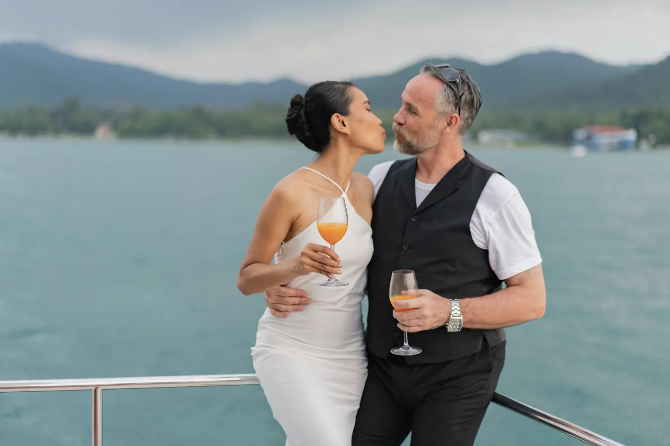 Couple in formal wear sharing a kiss on a boat, holding orange cocktails with a lake and mountains behind