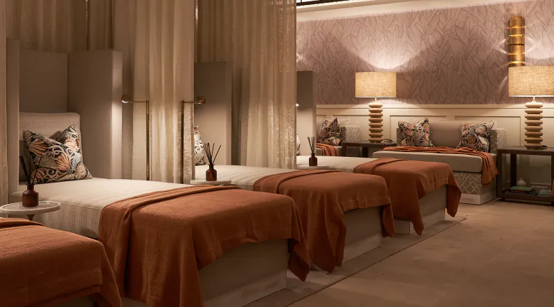 Luxury hotel spa with three massage beds draped in rust-orange linens, patterned pillows, and warm ambient lighting