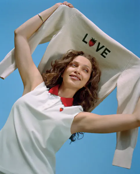 Woman in white polo holding up cream sweatshirt with 'LOVE' and strawberry graphic against blue sky