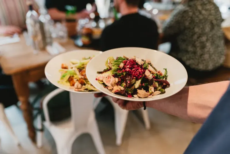 Hand holding two white bowls of fresh salad with greens, chicken, and pomegranate seeds in a cafe