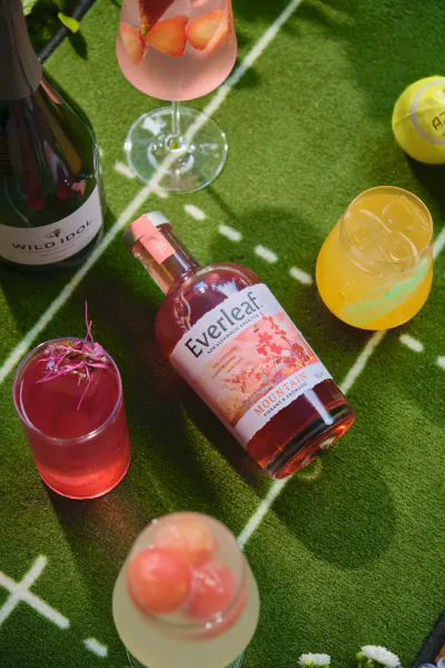 Assortment of colorful cocktails and a bottle of Everleaf Mountain Non-Alcoholic Aperitif on green grass with tennis court lines, next to a Wild Idol bottle and tennis ball