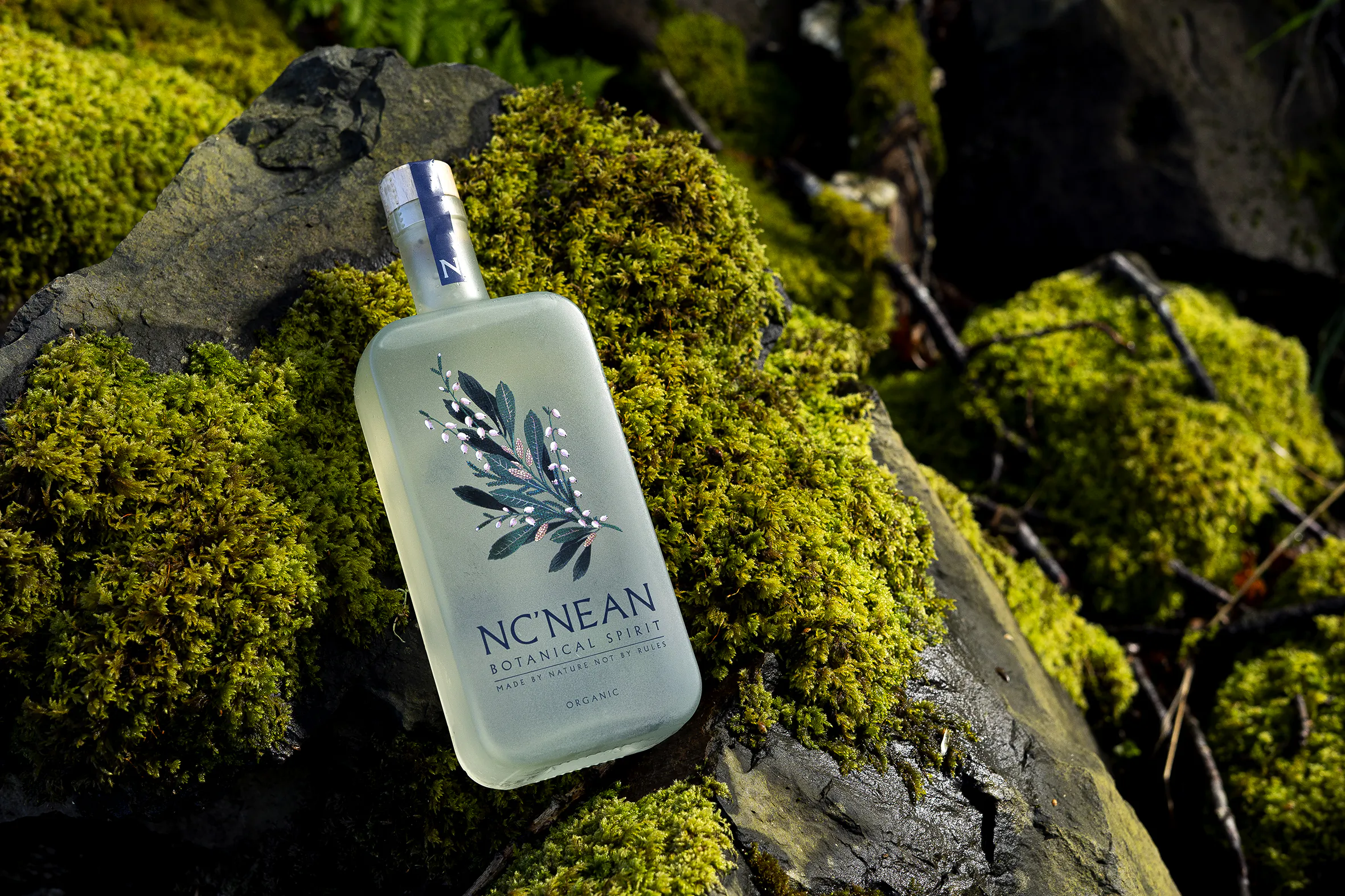 Nc'nean Botanical Spirit bottle resting on moss-covered rocks in a natural outdoor setting