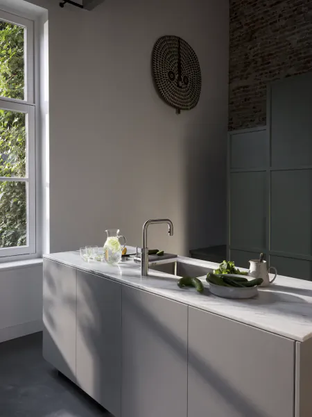 Modern kitchen with white cabinets, marble countertop, chrome Quooker tap, and fresh vegetables near a sunlit window