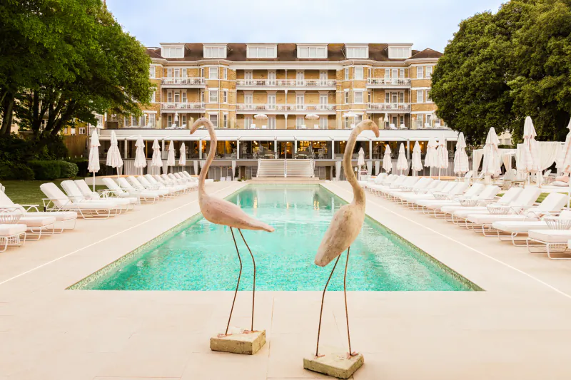 Two pink flamingo statues stand beside a long turquoise pool at The Nici Bournemouth, with white lounge chairs, umbrellas, and a grand yellow hotel behind