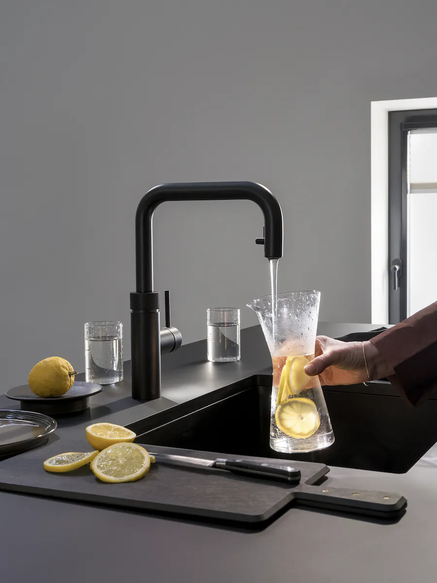 Hand filling glass pitcher with water and lemon slices from black Quooker faucet in modern kitchen