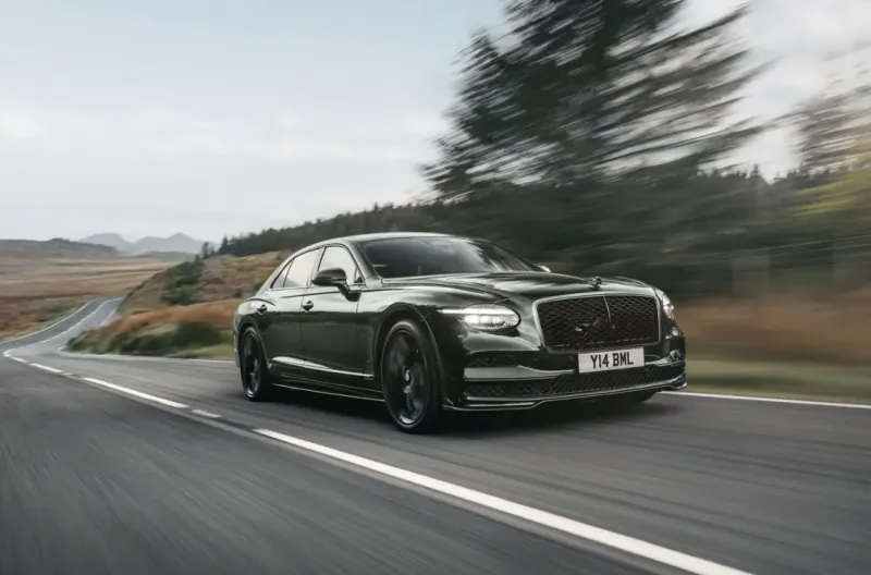 Dark green Bentley Flying Spur sedan driving fast on a winding rural road with motion blur