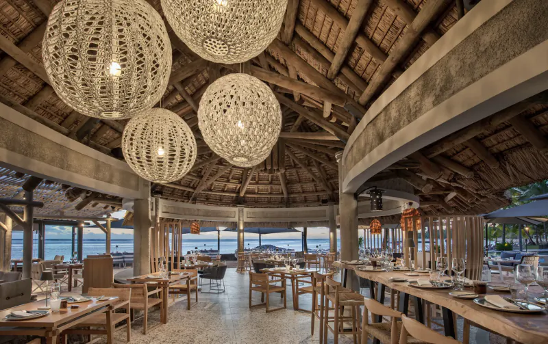 Elegant open-air beachfront restaurant with woven pendant lights, wooden tables set for dining, and ocean view at sunset