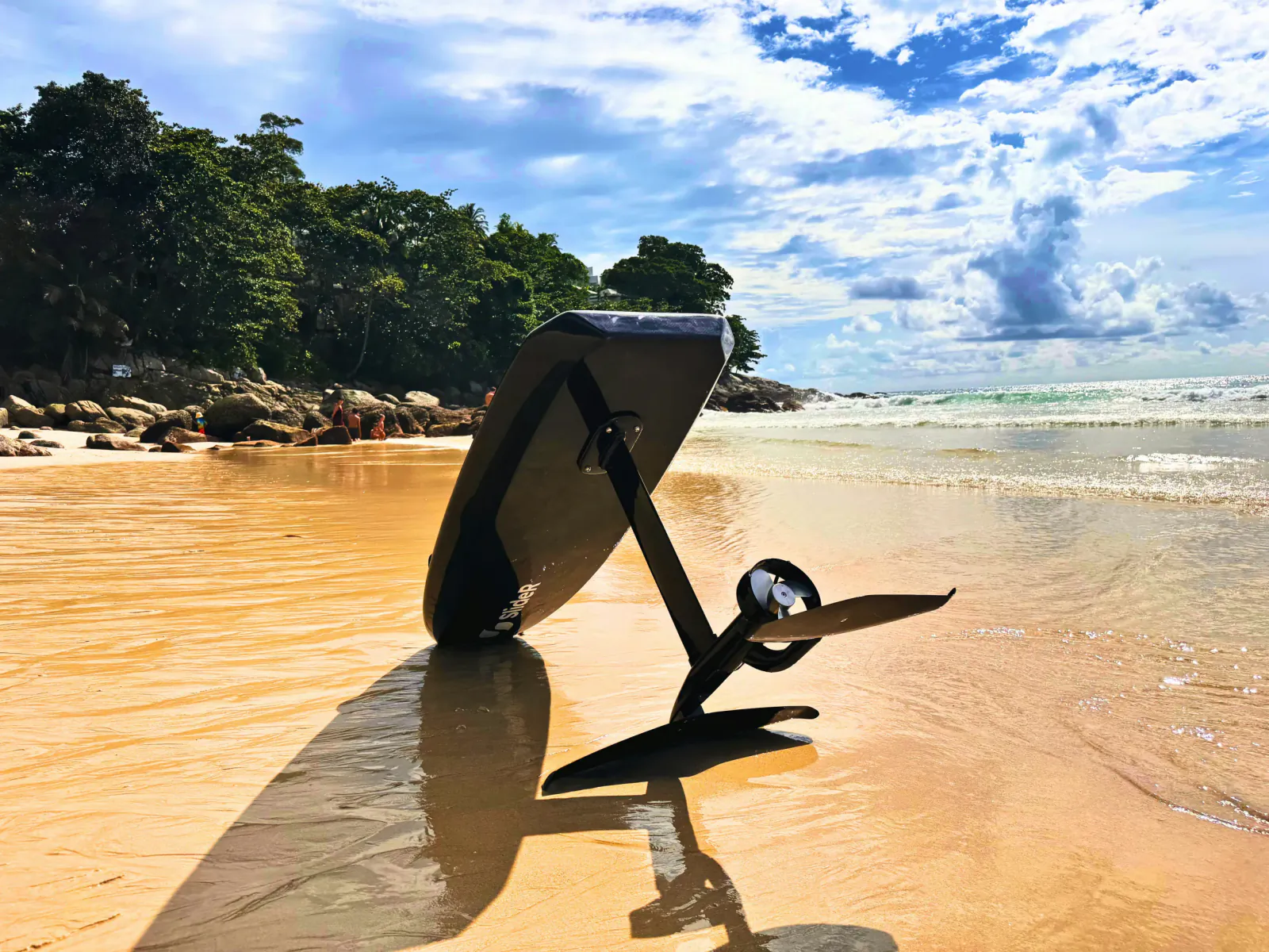 Electric foil surfboard standing upright on a tropical sandy beach with ocean waves and green hills in the background