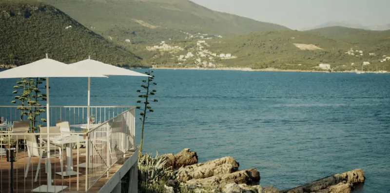 Outdoor terrace with white tables and umbrellas overlooking calm blue sea and green hills