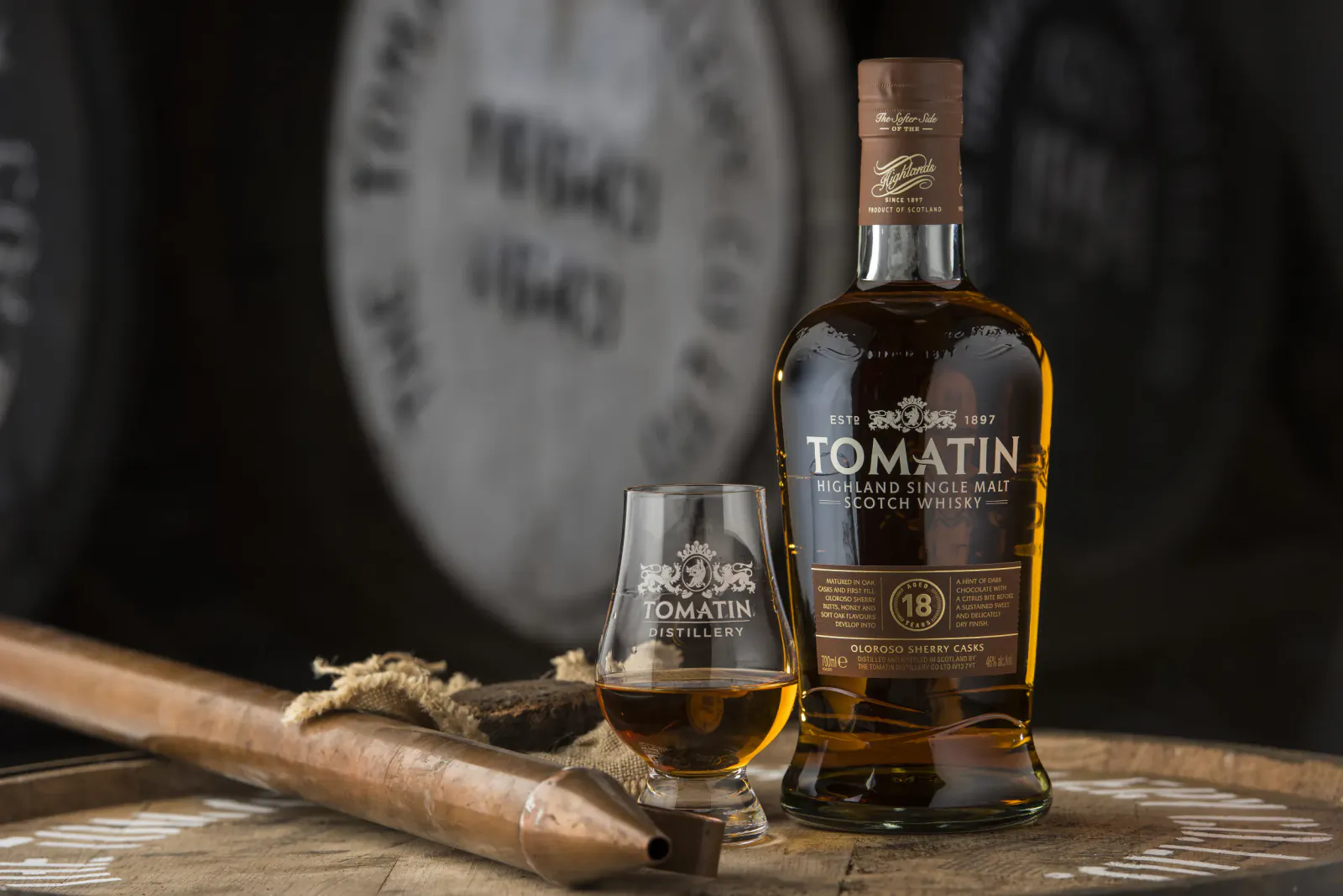 Tomatin 18 Year Old Highland single malt Scotch whisky bottle and tasting glass on wooden barrel with distillery barrels in background