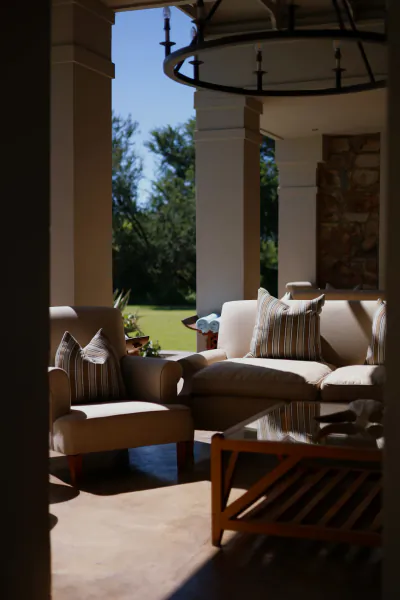 Sunlit patio with beige sofa, striped pillows, armchair, wooden coffee table, and view of green lawn and trees