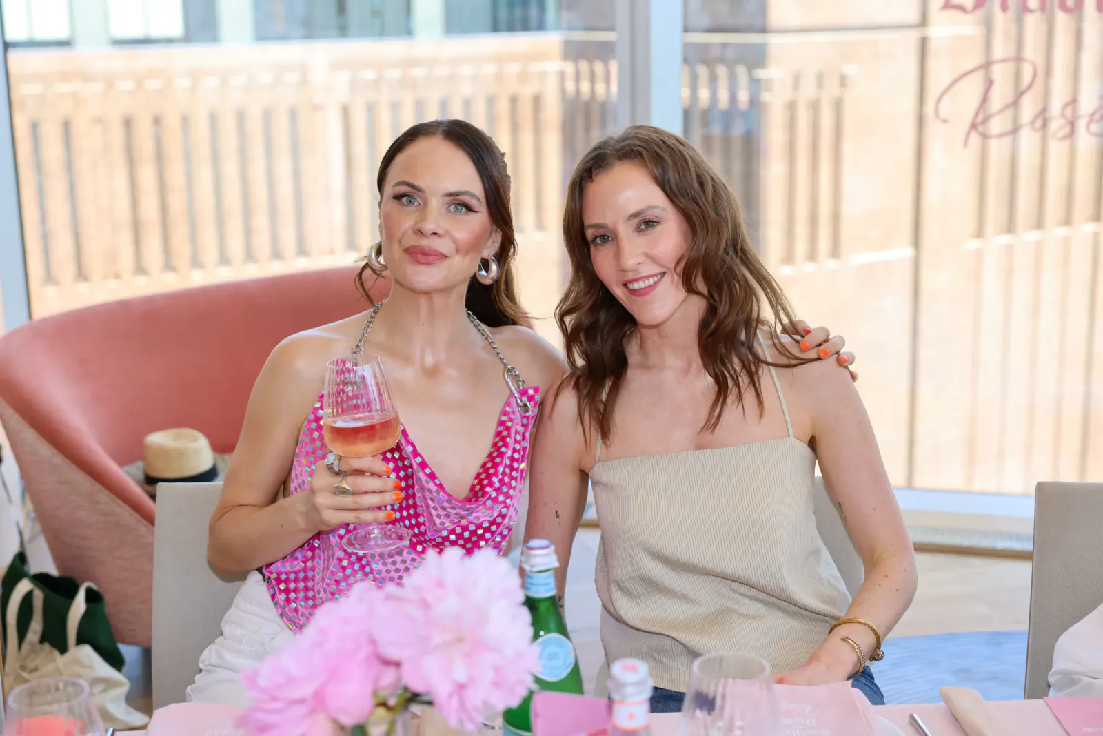Two smiling women pose at a table with pink roses and rosé wine; one in a pink polka-dot top holds a glass, the other in beige has an arm around her, with 'Casillero del Diablo Rosé' visible behind
