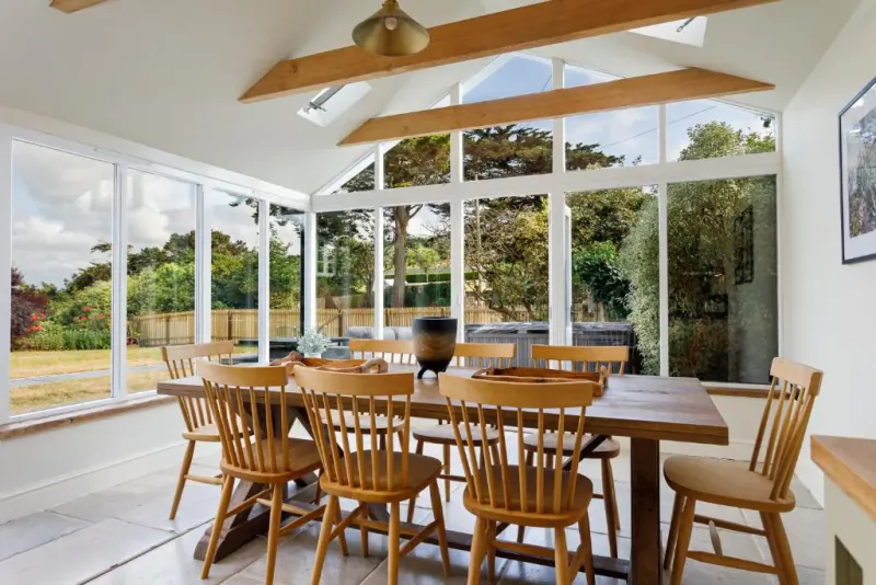 Bright conservatory dining room with large wooden table, spindle-back chairs, and floor-to-ceiling windows overlooking trees and garden