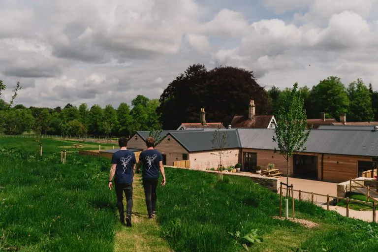 Two men in matching blue t-shirts walking across a grassy field toward modern barn-style buildings with a large tree in the background