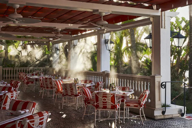Elegant outdoor patio at The Oyster Box with red and white striped chairs, set tables, ceiling fans, and lush palm trees in sunlight