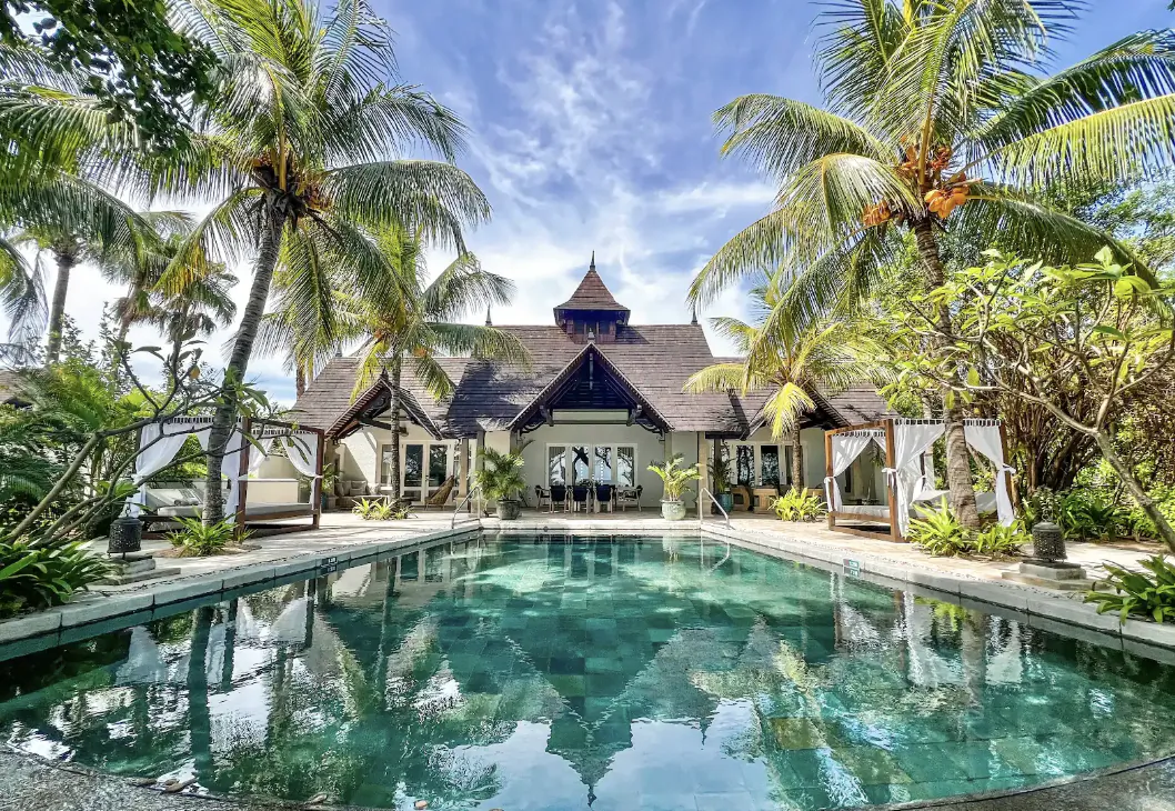 Luxury resort villa with thatched roof reflected in a turquoise pool, surrounded by palm trees under a blue sky