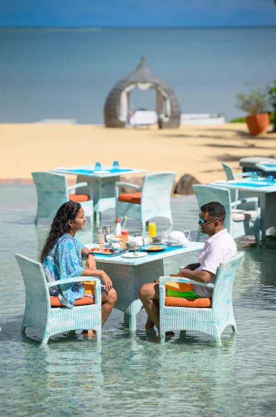Couple dining at a table in shallow water at a beachfront resort with ocean and thatched gazebo in background