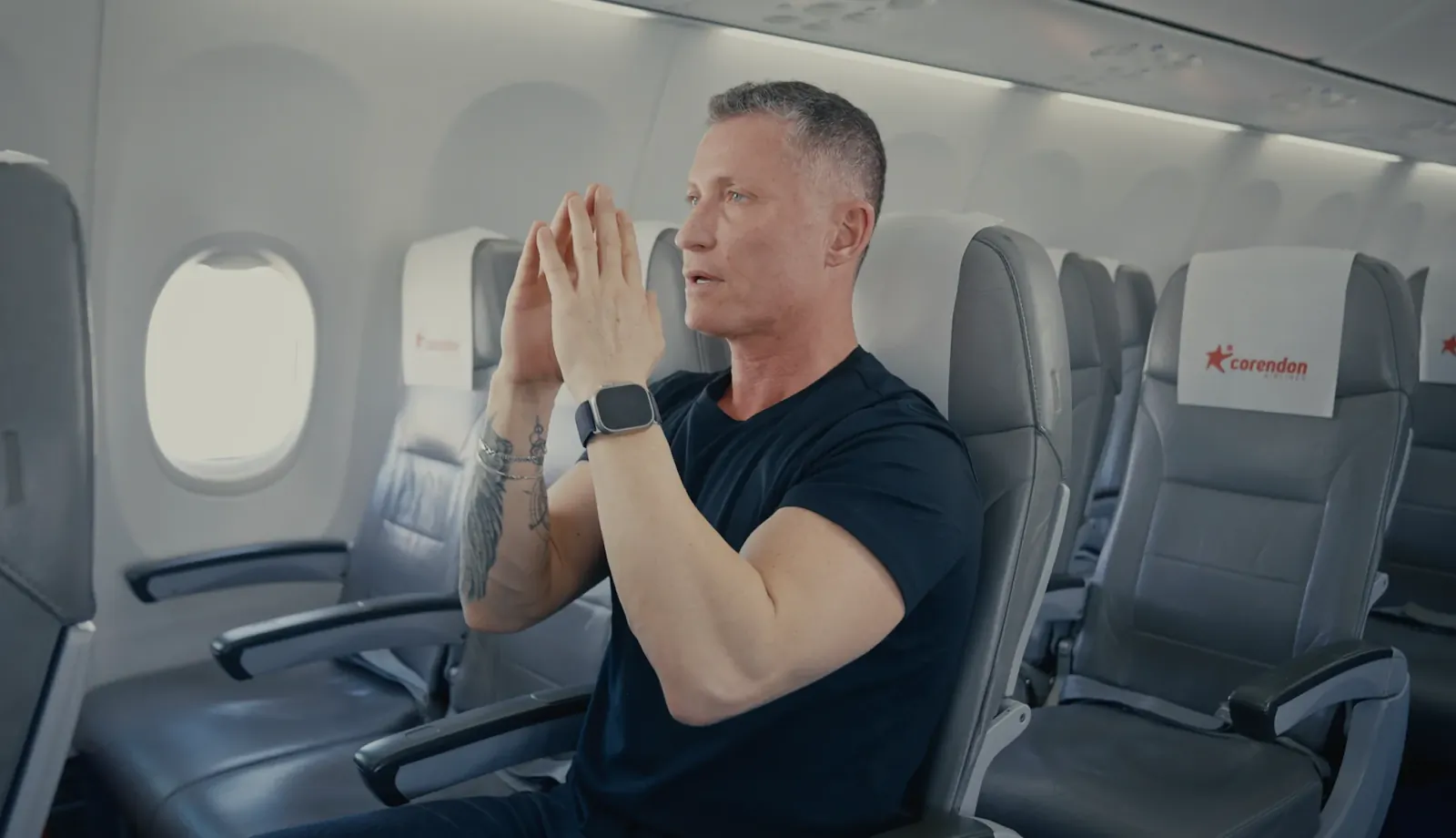 Man in black t-shirt seated on Corendon airplane, hands pressed together in a stretch