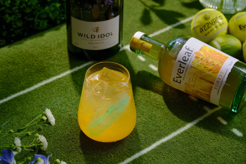 Yellow cocktail in ice-filled glass with green streak next to Everleaf bottle on green turf with tennis balls and Wild Idol bottle