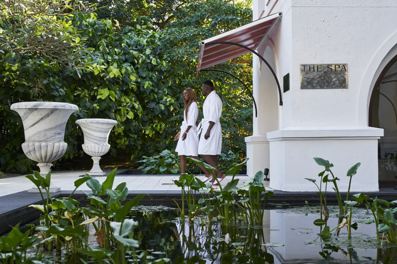 Couple in white robes walking past The Spa sign at a luxury resort with pond and greenery