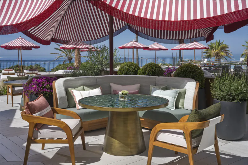 Outdoor patio with curved sofa, wooden chairs, round table under red and white striped awning overlooking the sea