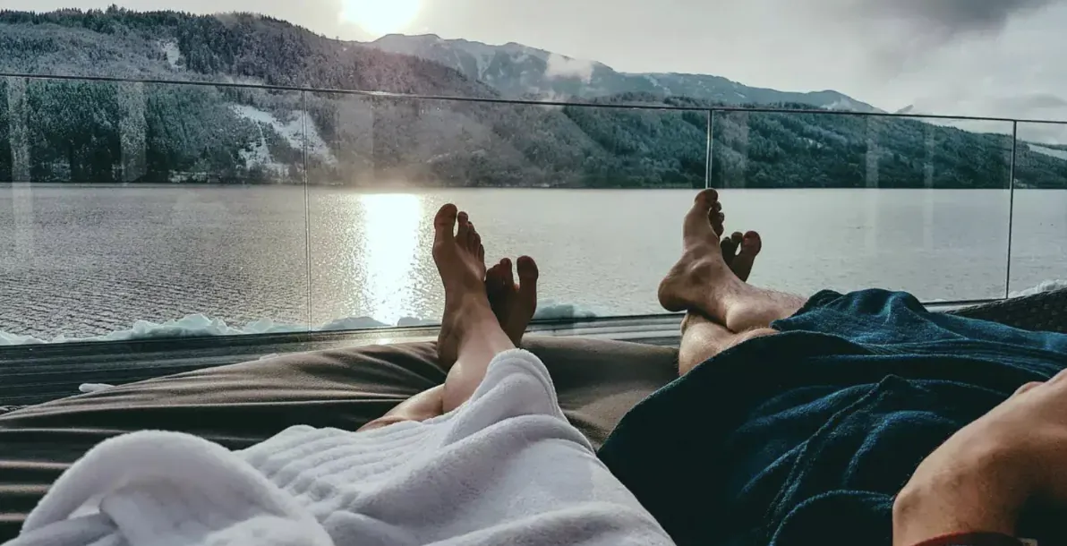 Two pairs of bare feet rest on a balcony overlooking a calm mountain lake at sunrise