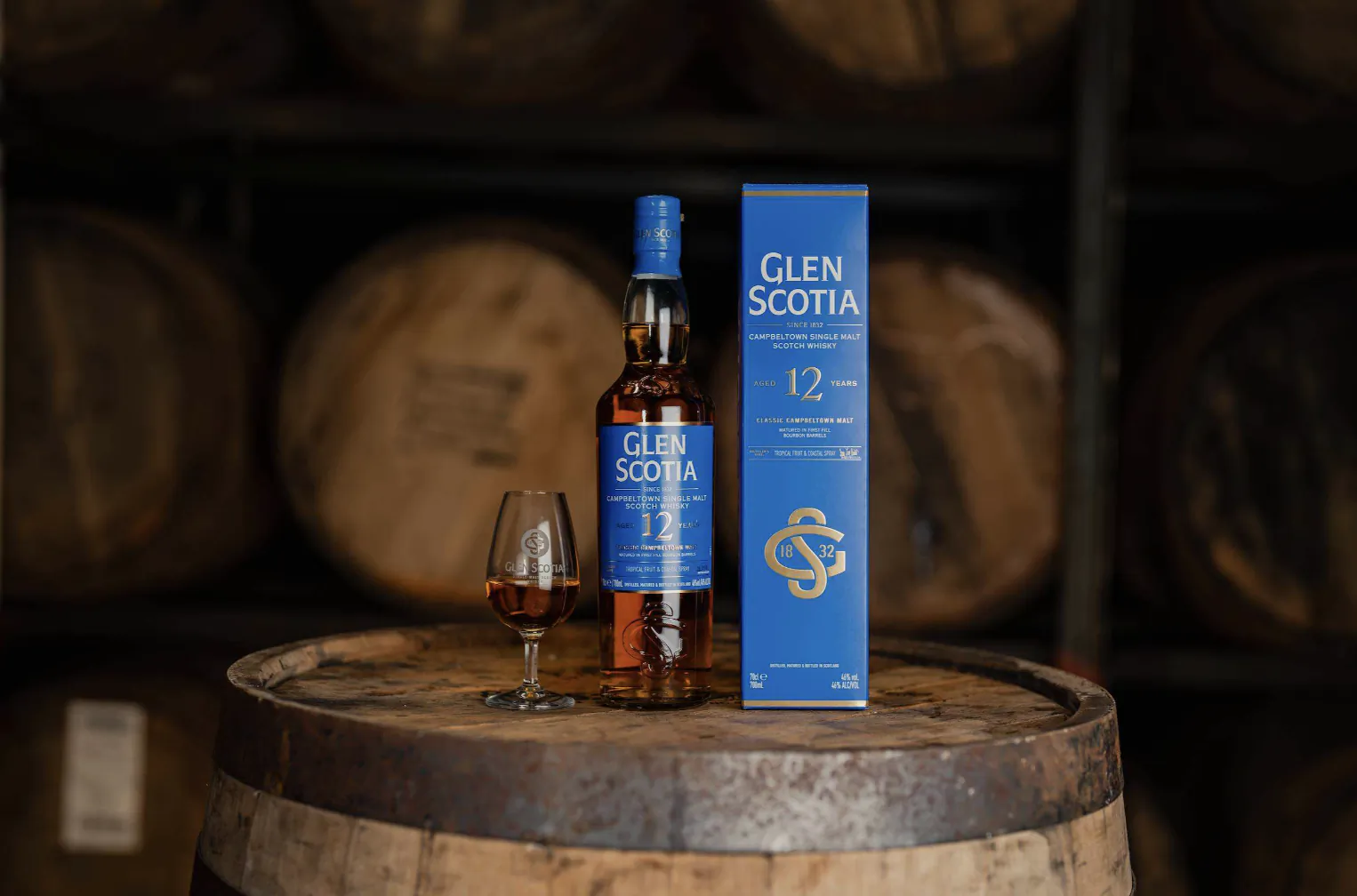 Glen Scotia 12 Year Old whisky bottle and box on a wooden barrel with a glass of whisky, surrounded by barrels