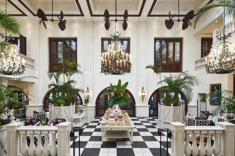 Luxurious hotel lobby with black-and-white checkered floor, grand chandeliers, palm plants, and a long buffet table set with food