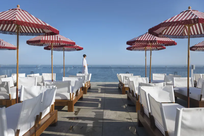 Man in white standing on sunny seaside terrace lined with red-and-white striped umbrellas and rows of white lounge chairs overlooking the ocean