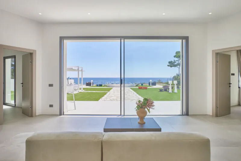 Bright modern living room with large sliding glass doors framing a sea view, green lawn, and stone path to the ocean