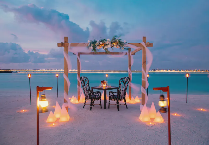 Romantic beach dinner setup under a wooden arch with lanterns and ocean view at sunset