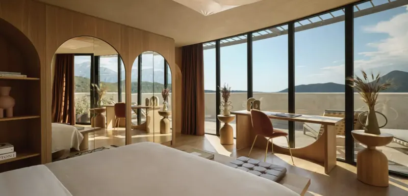 Modern hotel room with large windows overlooking mountains and sea, featuring wooden arched mirrors, minimalist furniture, and natural light