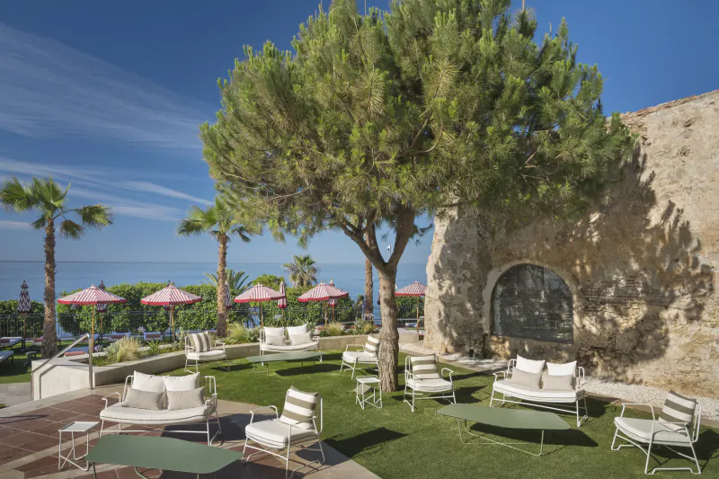 Luxury outdoor lounge with white lounge chairs and green tables on grass beside a stone wall, overlooking the sea under a large pine tree