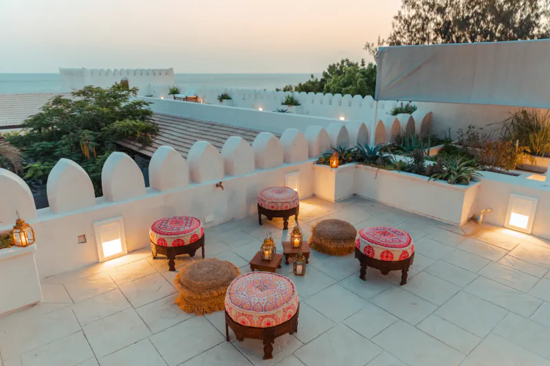 Rooftop terrace at dusk with colorful patterned ottomans, woven poufs, lanterns, and ocean view beyond white crenellated walls