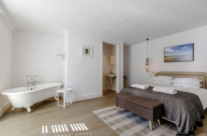 Modern hotel bedroom with freestanding white bathtub, double bed with grey throw, and open doorway to en-suite