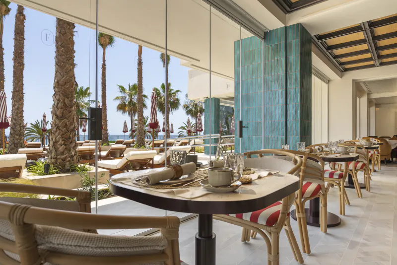 Modern restaurant interior with tables set for dining, large windows overlooking palm trees and sea view
