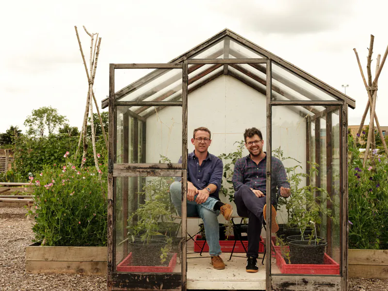 Two men sitting on chairs inside an open glass greenhouse surrounded by potted plants in a garden