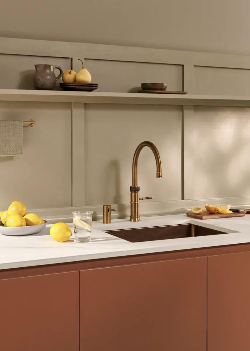 Modern kitchen with brushed gold Quooker faucet, white countertop, terracotta cabinets, bowl of lemons and glass of water