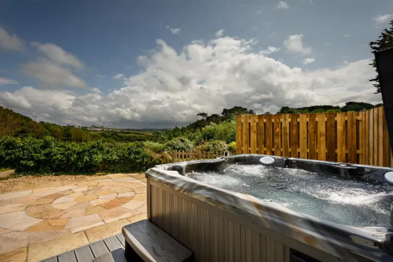 Hot tub bubbling on a patio with countryside hills and wooden fence under a cloudy sky